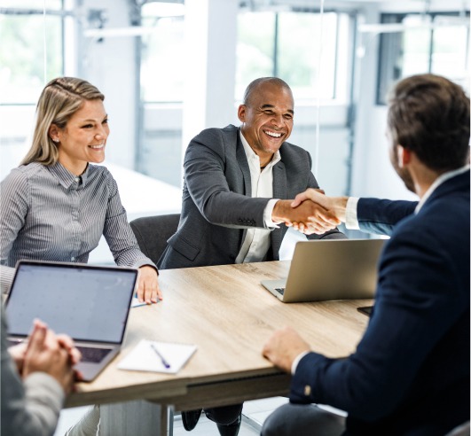 Two men shake hands across a conference table while a woman looks on with a smile on her face.