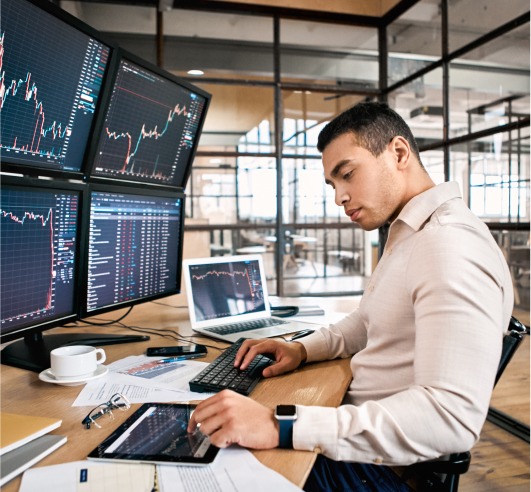 A man looks at his tablet while typing something on his computer. He has four monitors set up that are tracking some trends.