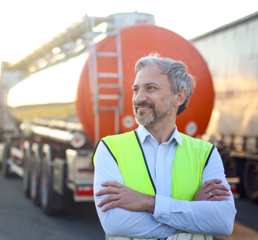 A man smiles looking off to the side while several tractor trailers are parked behind him.