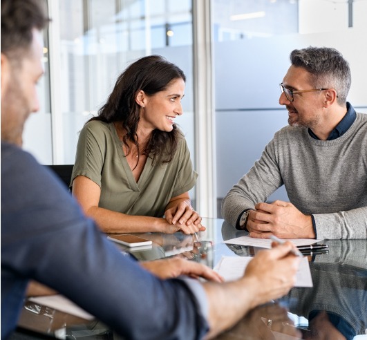 A woman smiles at a man at a conference table. In the forefront is another man that is writing on a piece of paper.